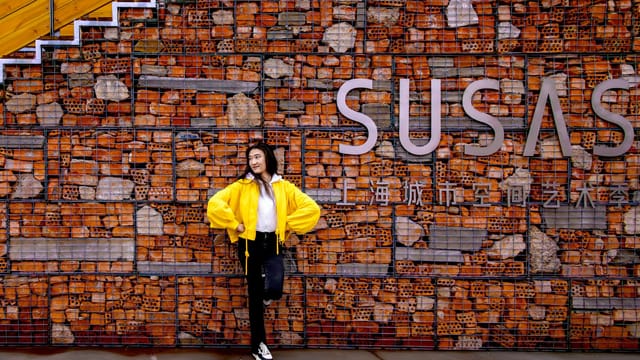 A young woman poses against a brick wall with SUSAS sign in Shanghai, China.