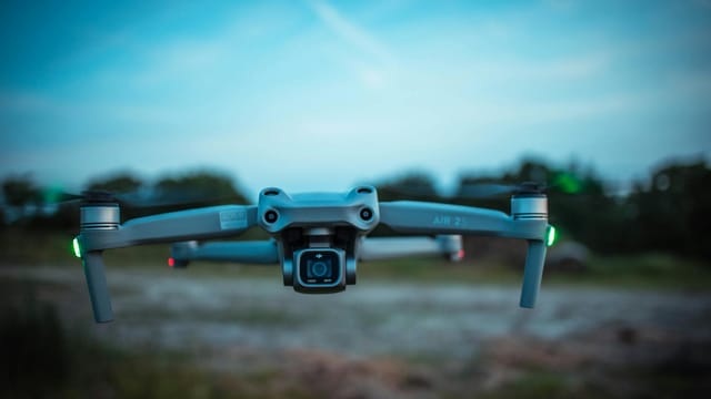 Close-up of a modern quadcopter drone with lights, hovering over a blurred outdoor background.
