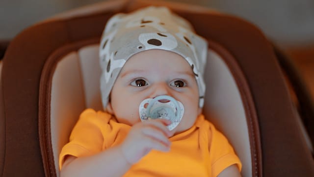 Adorable baby with beanie and pacifier in orange shirt, indoors.