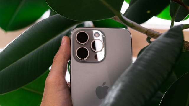 Hand holding a smartphone with triple camera lens against green leaves, showcasing modern design.