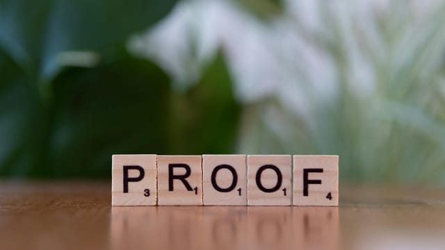 Wooden Scrabble tiles spelling 'PROOF' on a wooden table with green background.