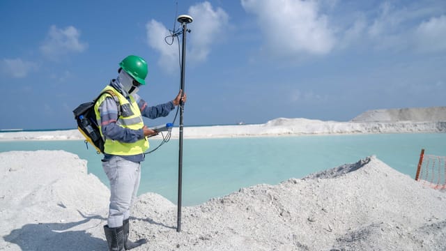 Surveyor in protective gear conducting measurements by a remote beach with GPS equipment.