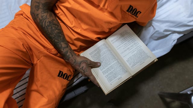 Tattooed inmate reading a book on a bed in a prison cell.