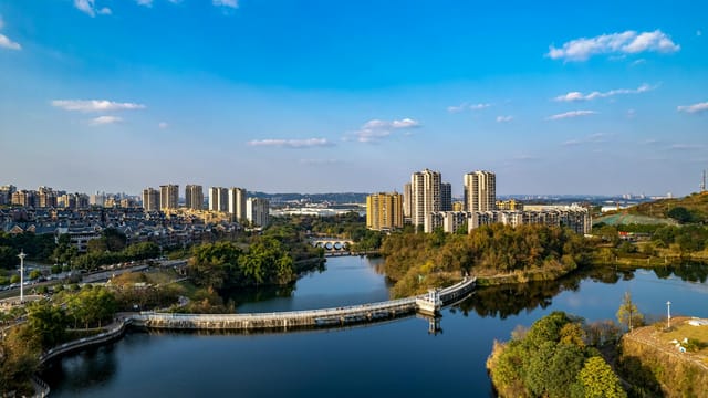 A panoramic aerial view of Zigong, showcasing urban buildings, lush greenery, and a tranquil river under a blue sky.