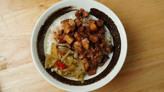 A savory bowl of braised pork with rice and vegetables on a wooden table.
