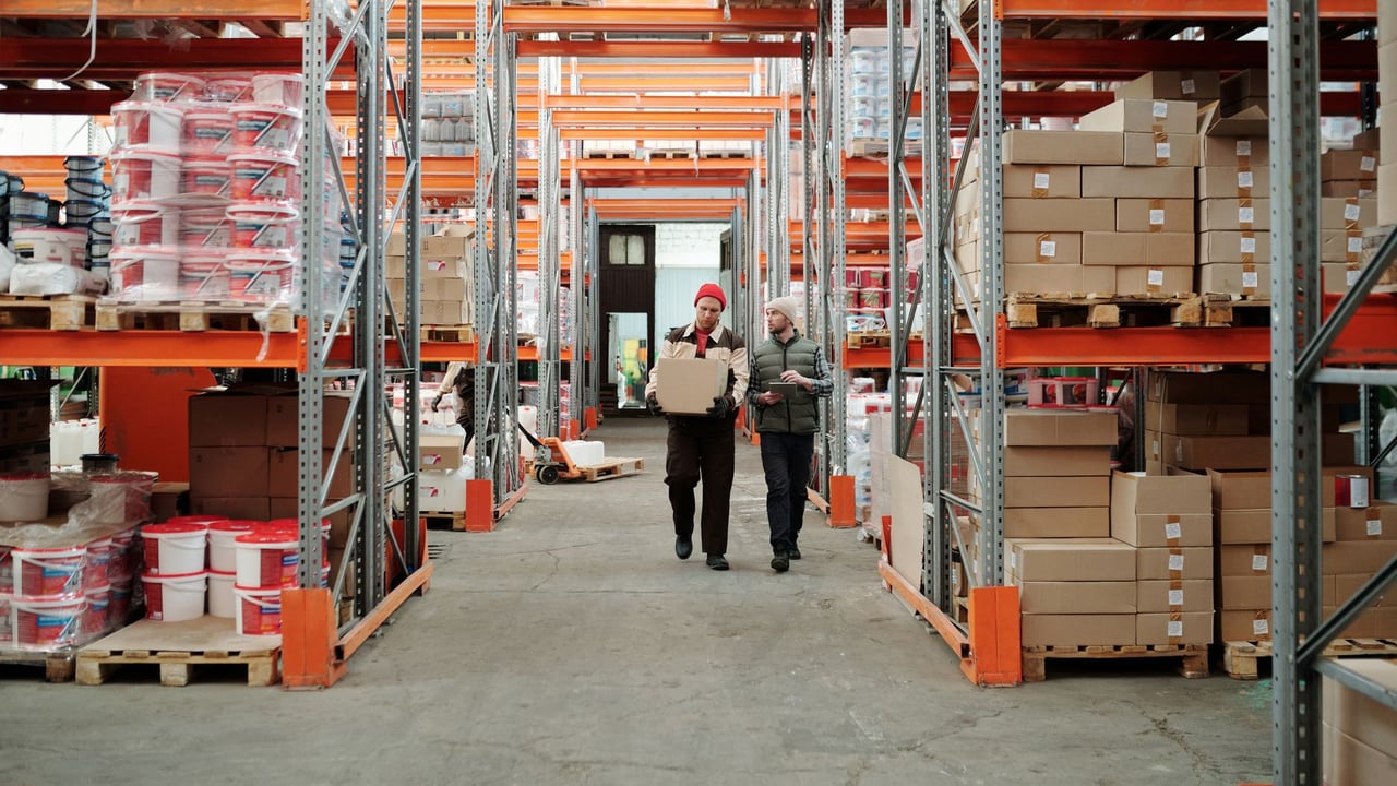 Illustrative image: Two workers handle a package in a spacious warehouse surrounded by shelves stocked with boxes and products.