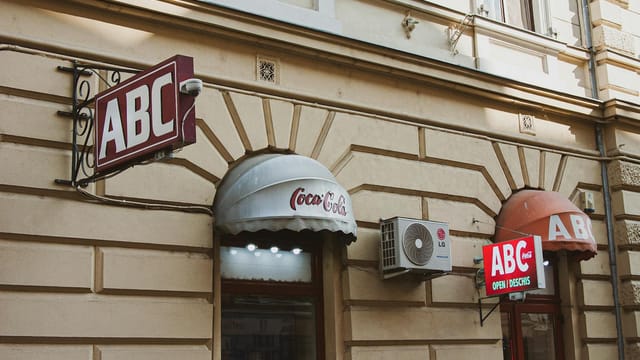 A vintage-style shopfront with multiple ABC signs and a Coca-Cola awning in an urban setting.