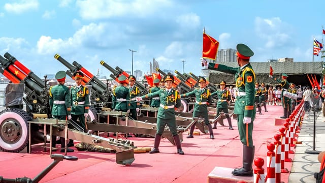 Military parade with soldiers and artillery cannons on display, featuring colorful uniforms and flags.