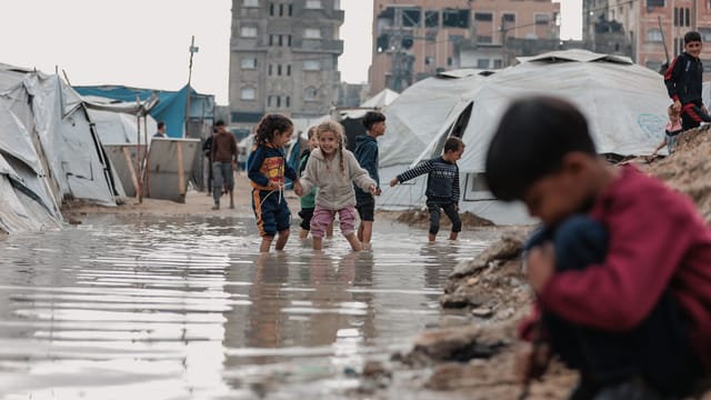 Children splash in water amidst refugee tents in Gaza, capturing resilience and joy.