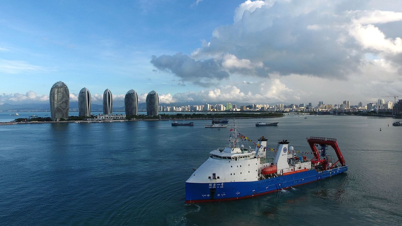 Aerial shot of a research vessel sailing near Phoenix Island, Sanya, with modern skyscrapers in the background.