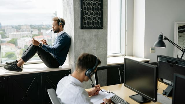 Two call center agents working in a modern office, one seated at a computer and another using a tablet by the window.
