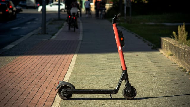 A Voi e-scooter parked on a sunlit city sidewalk with a blurred urban background.