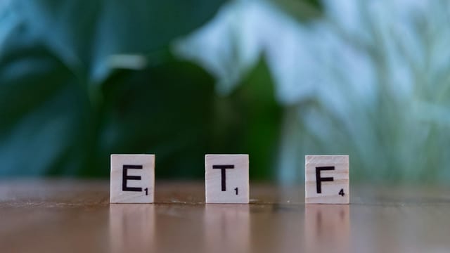 Scrabble tiles spelling ETF on a wooden surface with blurred green background.