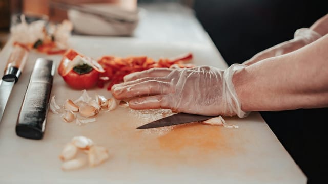 Gloved hands chopping garlic and tomatoes on a cutting board in a kitchen.