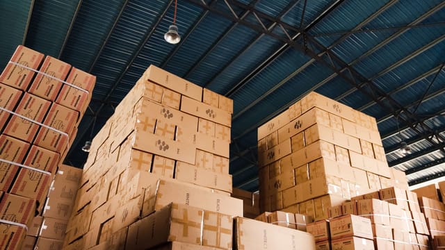 Illustrative image: High stacks of cardboard boxes organized in a warehouse with a blue metal ceiling.