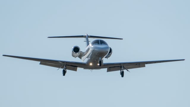 A sleek silver private jet approaches landing under a clear blue sky.