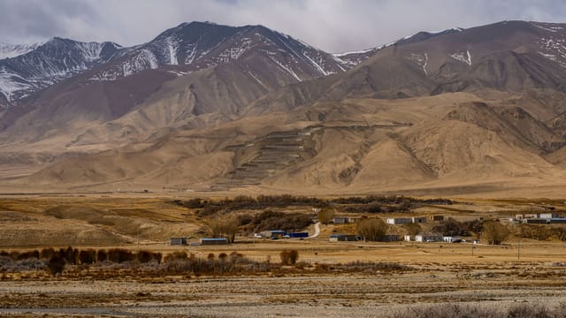 A vast desert landscape with distant snow-capped mountains and a small settlement in the foreground.
