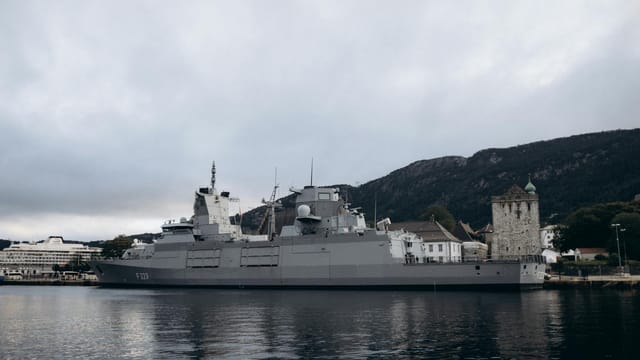 A military ship docks in Bergen, Norway harbor with historic buildings and mountains in the background.