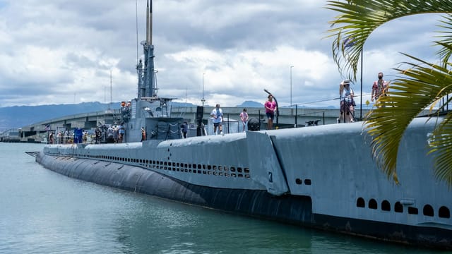 Tourists explore a historic submarine docked at Pearl Harbor, Hawaii under a cloudy sky.