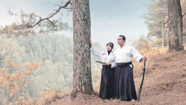 A martial arts couple in traditional attire with katanas, standing in a serene forest setting.
