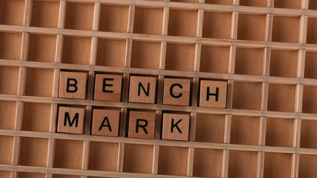 Wooden letter tiles spelling 'BENCHMARK' placed on a grid-like wooden background.