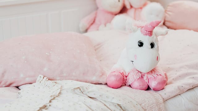 A soft unicorn plush toy sits on a pastel pink bed with pillows and blankets in a child's room.