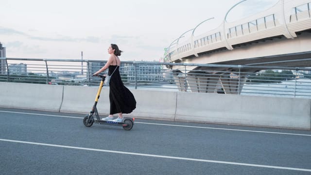 A woman enjoys a ride on an electric scooter across a modern urban bridge at sunset.