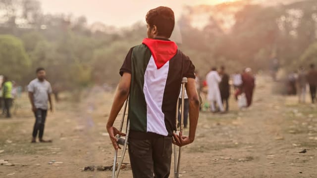 A young man wrapped in a Palestinian flag walks with crutches through a field in Bangladesh during the day.