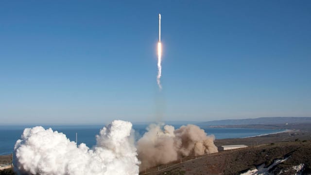 A rocket launches over the ocean, leaving behind smoke and fire trails. Scenic backdrop with clear skies.