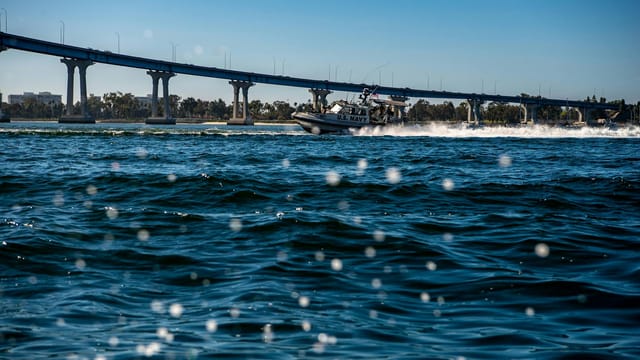 Navy patrol boat speeding under a bridge, creating waves and ripples in the water.