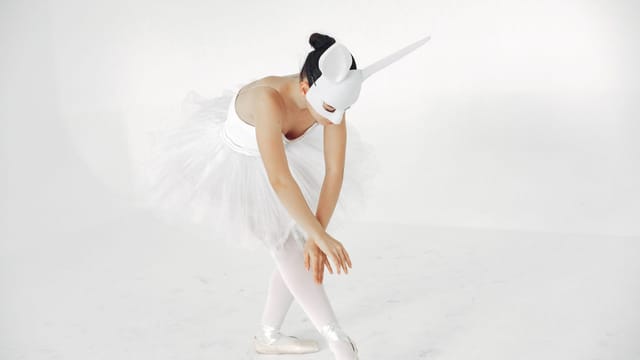 A graceful ballerina with a unicorn mask poses in a white tutu against a plain backdrop.