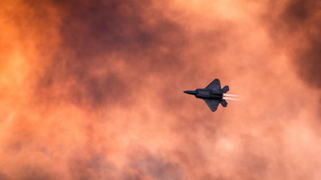 A fighter jet soars against a backdrop of vibrant, fiery clouds at sunset.