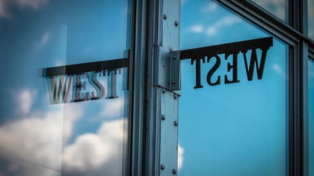 Glass window reflecting sky and clouds with 'West' sign in urban setting.
