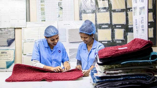 Two female workers in a textile factory inspecting red carpet samples on a table.