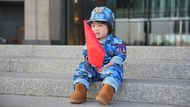 Young child in blue camouflage uniform sits holding a red flag on concrete steps.