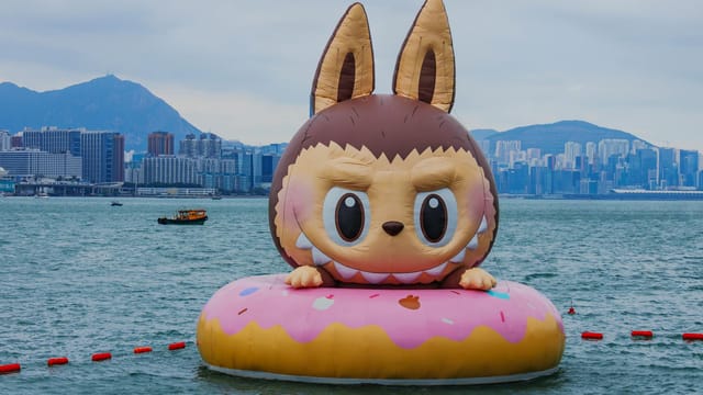 Large inflatable cartoon character on a pink float in Victoria Harbour with Hong Kong skyline in background.