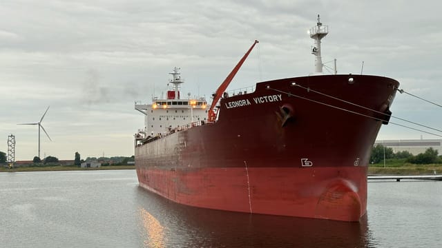 A large red cargo ship named Leonora Victory at a European dock with a wind turbine in the background.