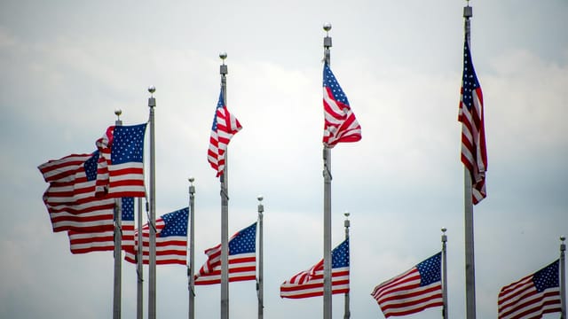 A row of American flags waving proudly at the base of the Washington Monument against a cloudy sky.