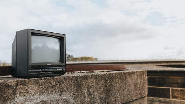 Small old fashioned black television placed on shabby concrete border with graffiti against cloudy sky in city on street with bridge in distance