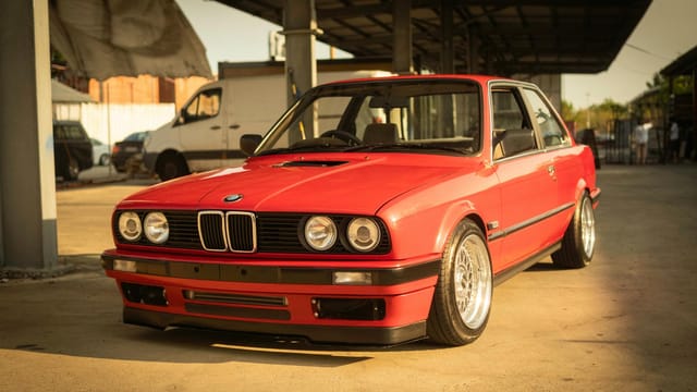 A classic red BMW E30 coupe parked in an industrial setting under warm sunlight.