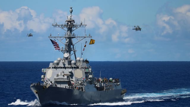 A US Navy destroyer navigates the ocean with helicopters in formation above.