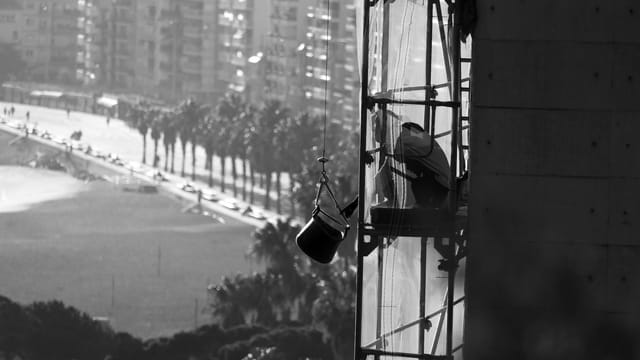 Black and white photo of a construction worker on scaffolding by a busy urban beachfront.