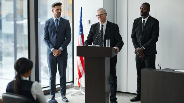 Officials delivering a political speech in a modern conference room with an American flag.
