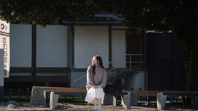 A young woman enjoying a sunny day on a bench under a tree in Japan.