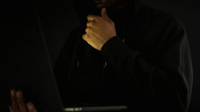 Crop contemplative male hacker in black hoodie watching netbook and touching chin thoughtfully in dark room