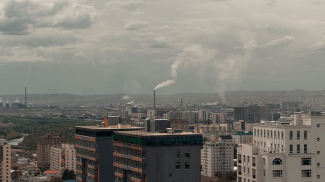 A cityscape featuring buildings and industrial smoke pollution under a cloudy sky.