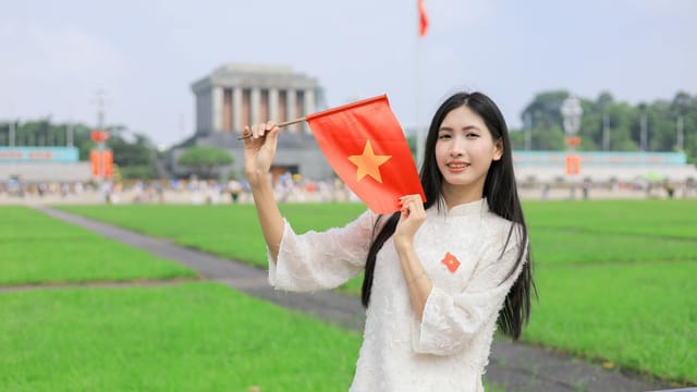 Young woman in traditional Ao Dai holding a Vietnamese flag in front of the Ho Chi Minh Mausoleum in Hanoi.