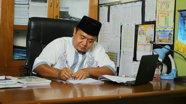 Indonesian educator writing at his desk, focused on paperwork in an office setting.
