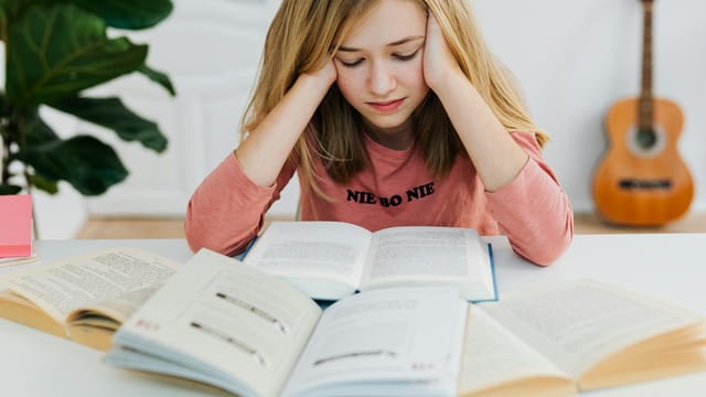A young woman in a pink sweater focused on reading books at a table indoors.