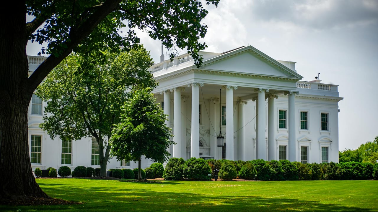 The White House framed by trees and greenery, in Washington, D.C., under a bright sky.